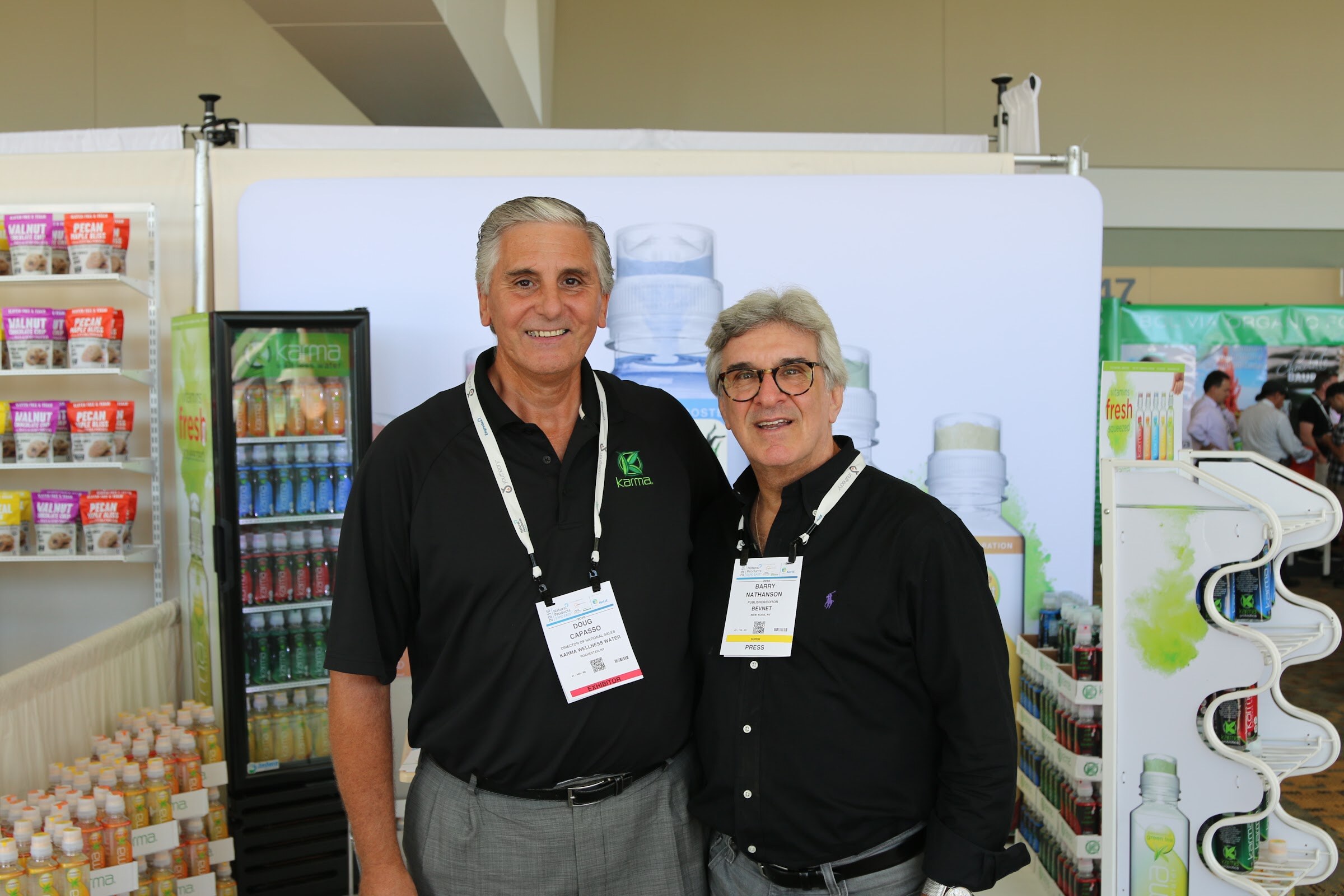 Two smiling men in black shirts and lanyards stand side by side in a booth with a glass-door cooler of drinks and product displays behind them.