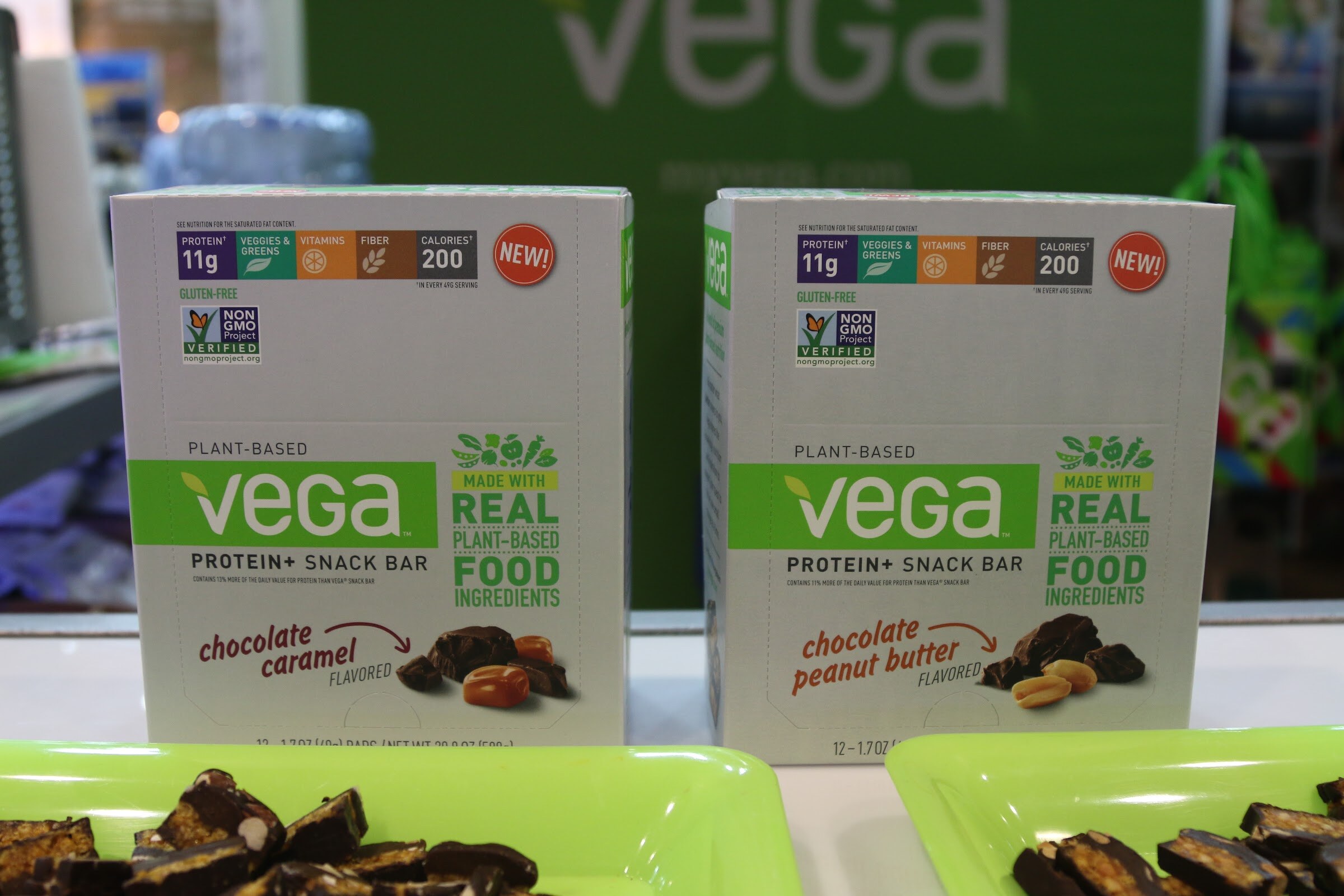 Two white Vega Protein+ Snack Bar boxes in front of a green Vega backdrop, with green trays of cut-up bars below.