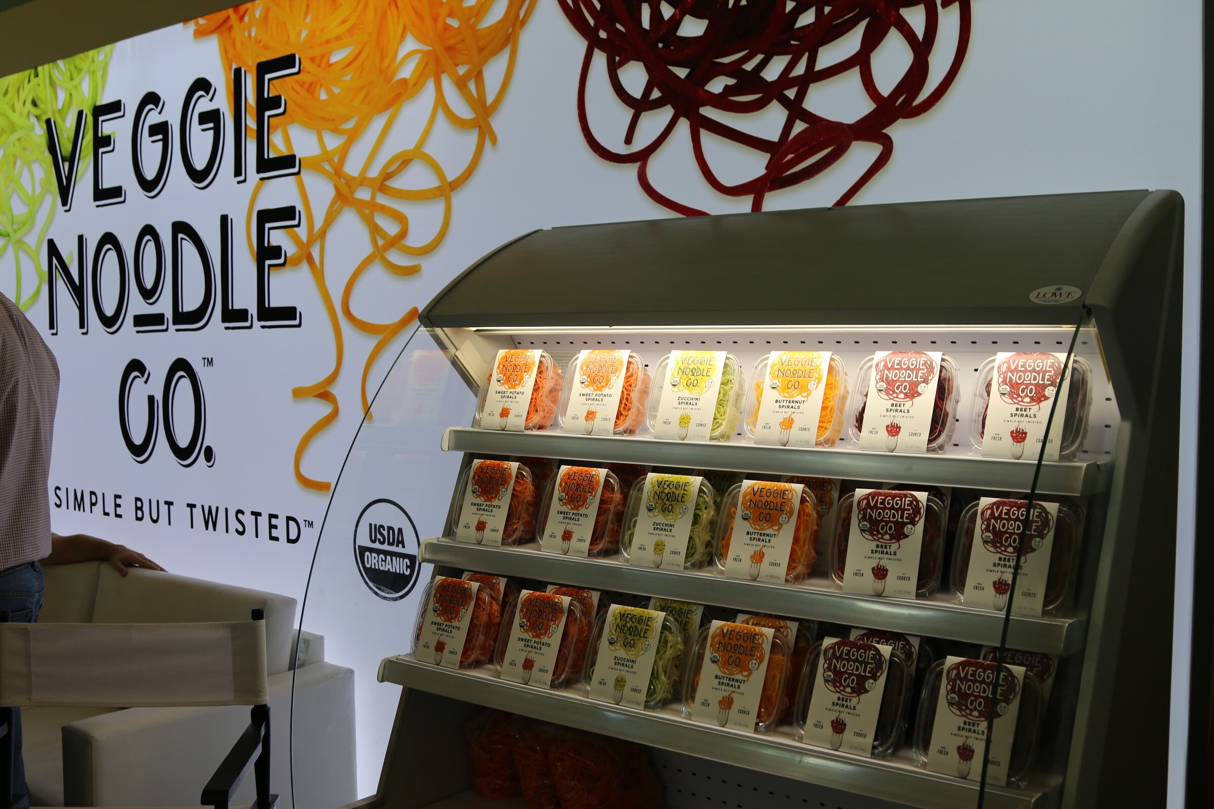 A white Veggie Noodle Co. sign with spiral illustrations above a lit open cooler stocked with rows of veggie spiral containers.