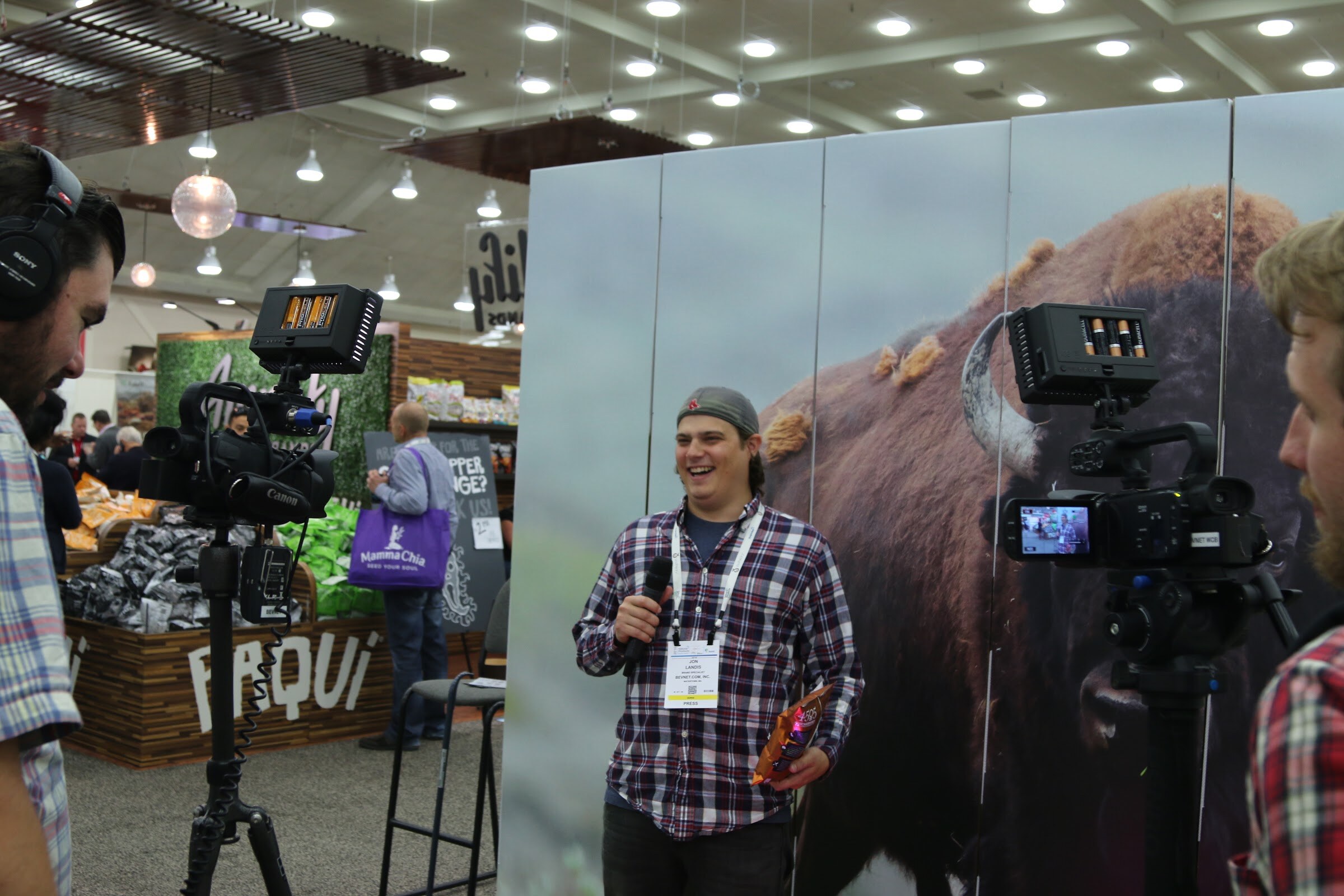 A man in a plaid shirt with a lanyard speaks into a microphone as camera crew film him, in front of a giant bison photo and a wooden Paqui sign.