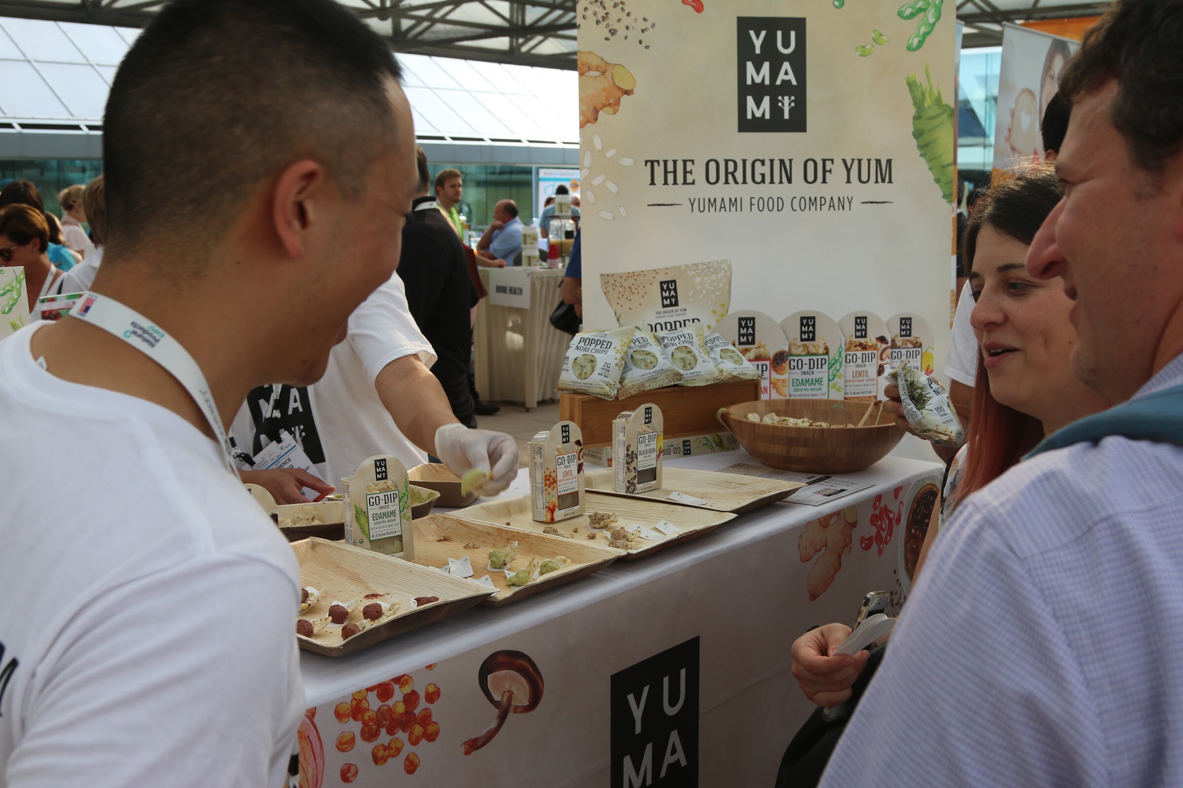 A man in a lanyard smiles toward visitors at a Yumami booth table covered with sample plates and Go-Dip packs under a Yumami banner.