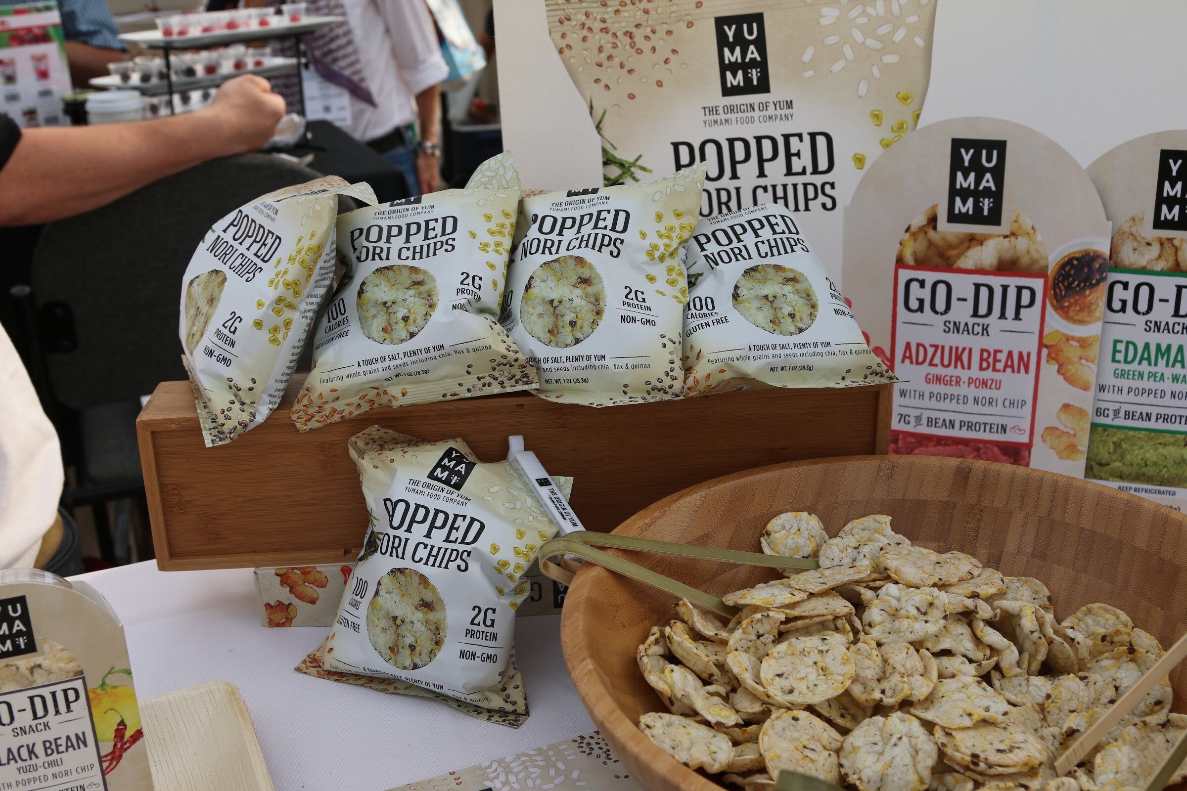 A wooden bowl of round popped nori chips next to bagged Yumami popped nori chips and Go-Dip Adzuki Bean snack packs.