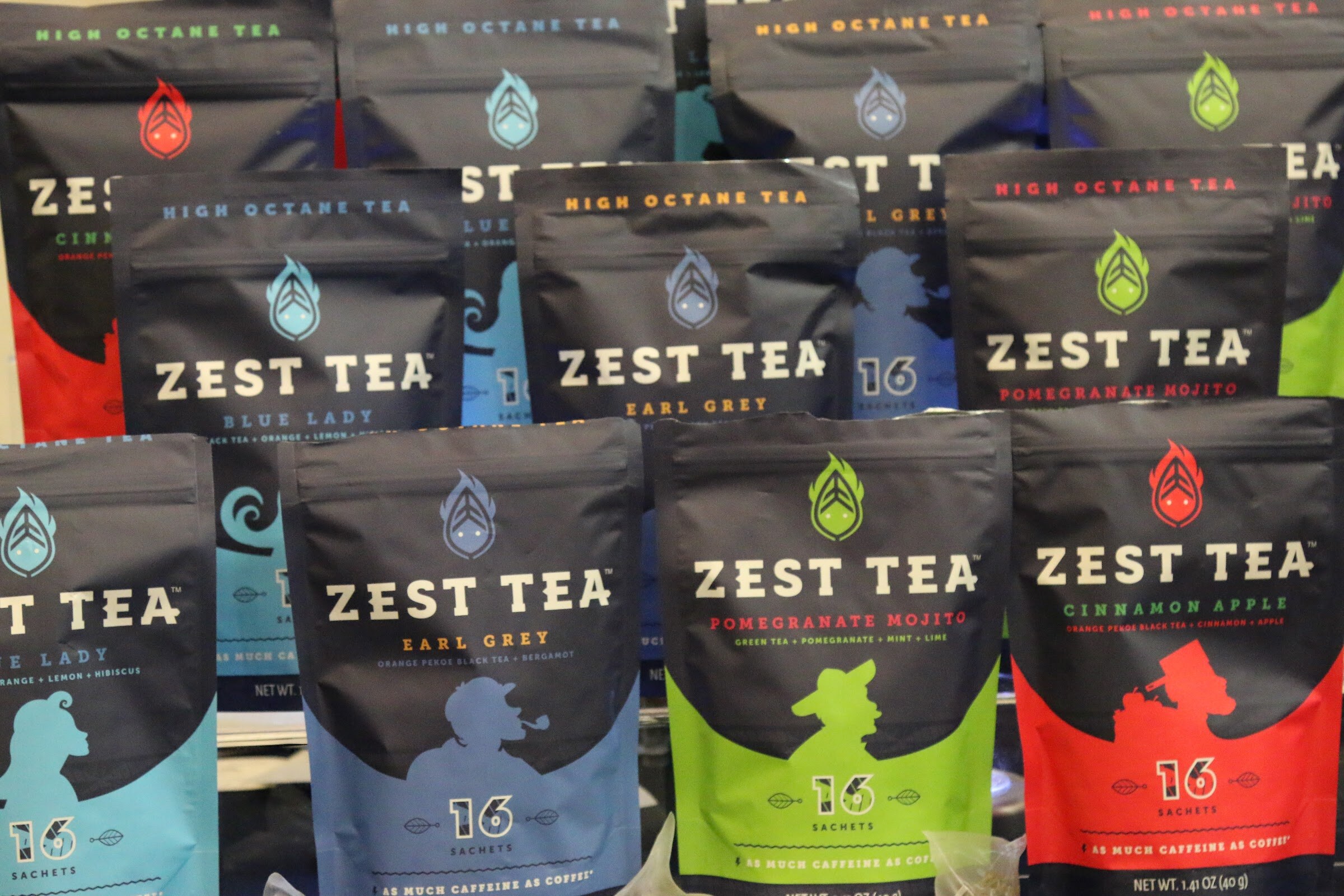 Rows of dark Zest Tea pouches with blue, green and red accent colors stacked in a display.