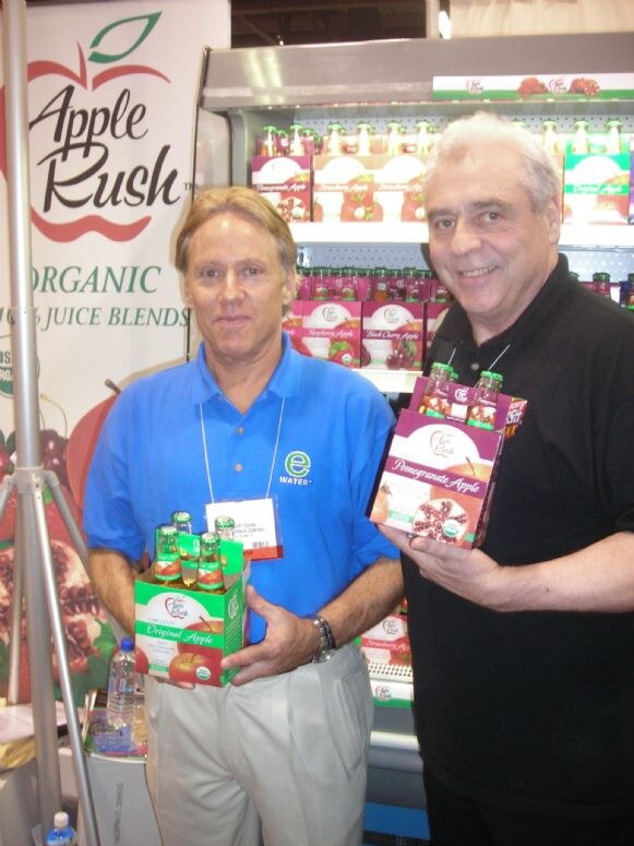 Two men hold four-packs of Apple Rush organic juice, Original Apple and Pomegranate Apple, in front of a refrigerated display. One wears a blue e2o Water shirt.