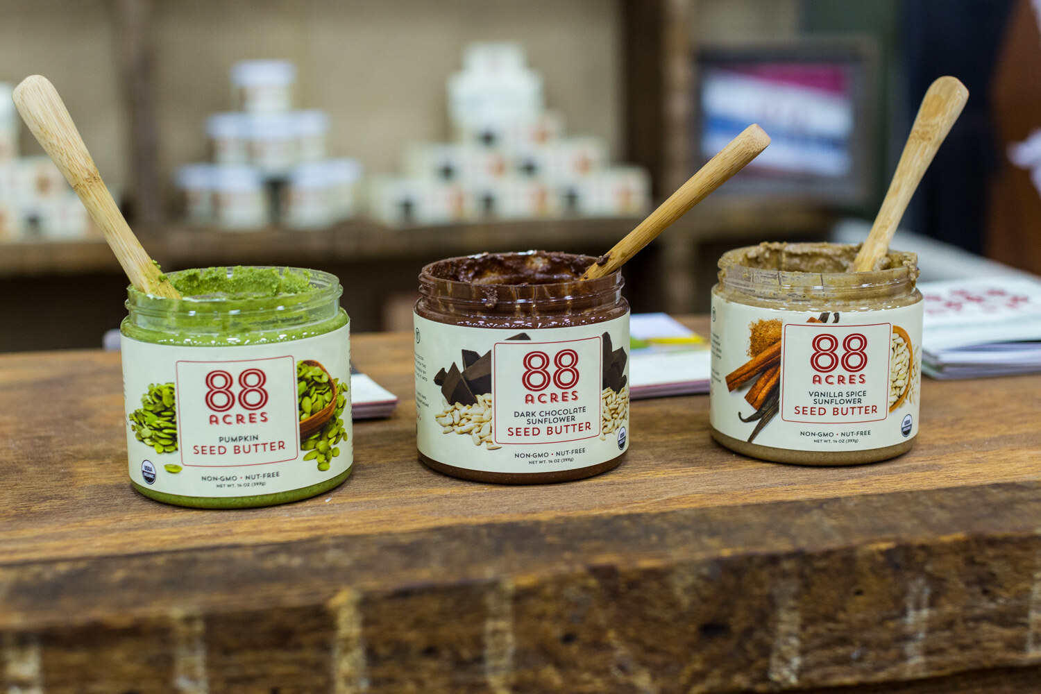Three 88 Acres seed butter jars with wooden spoons on a rustic wooden counter with stacked jars blurred behind.