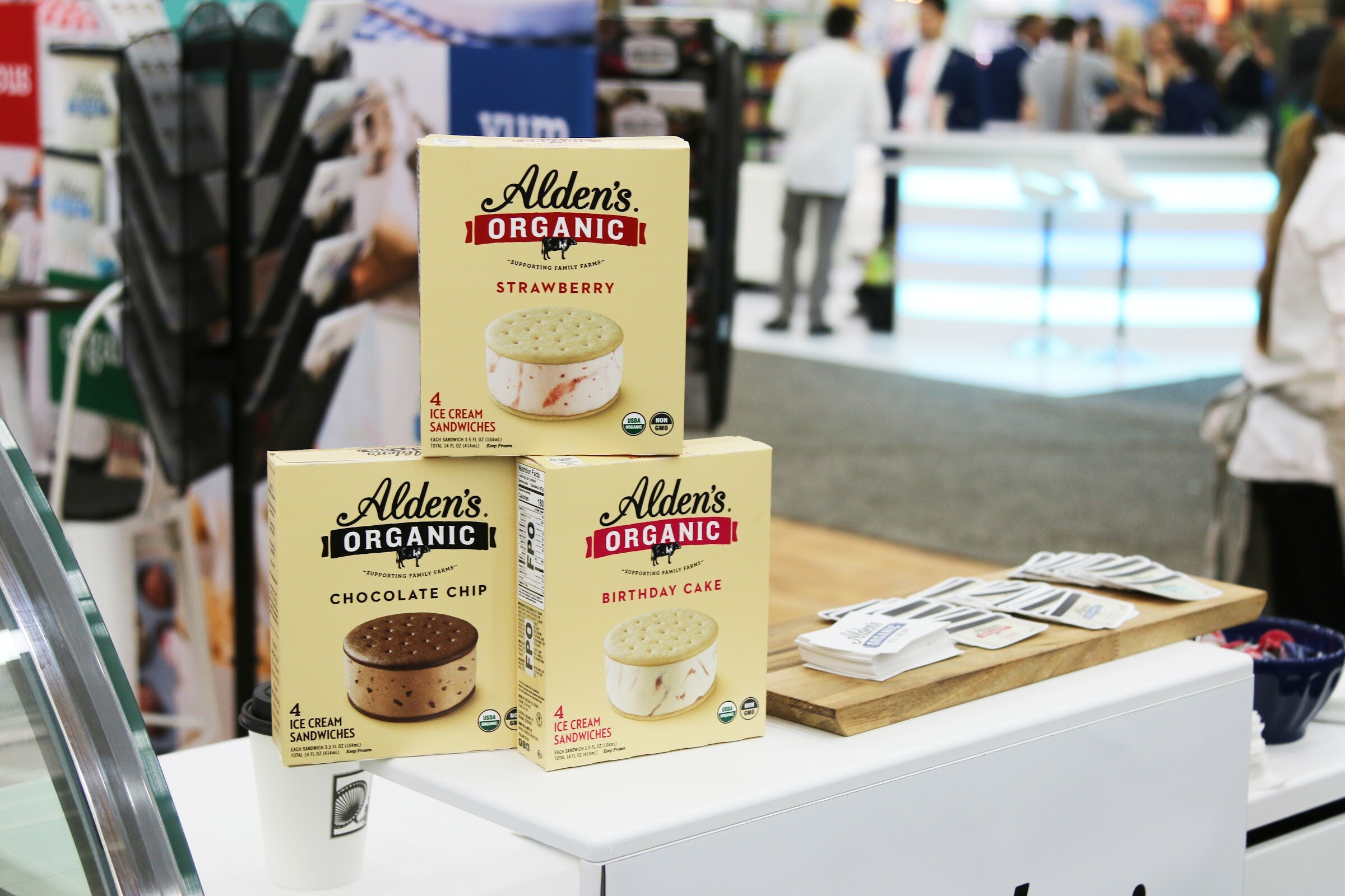 Three boxes of Alden's Organic ice cream sandwiches on a white counter with attendees and booths blurred in the background.