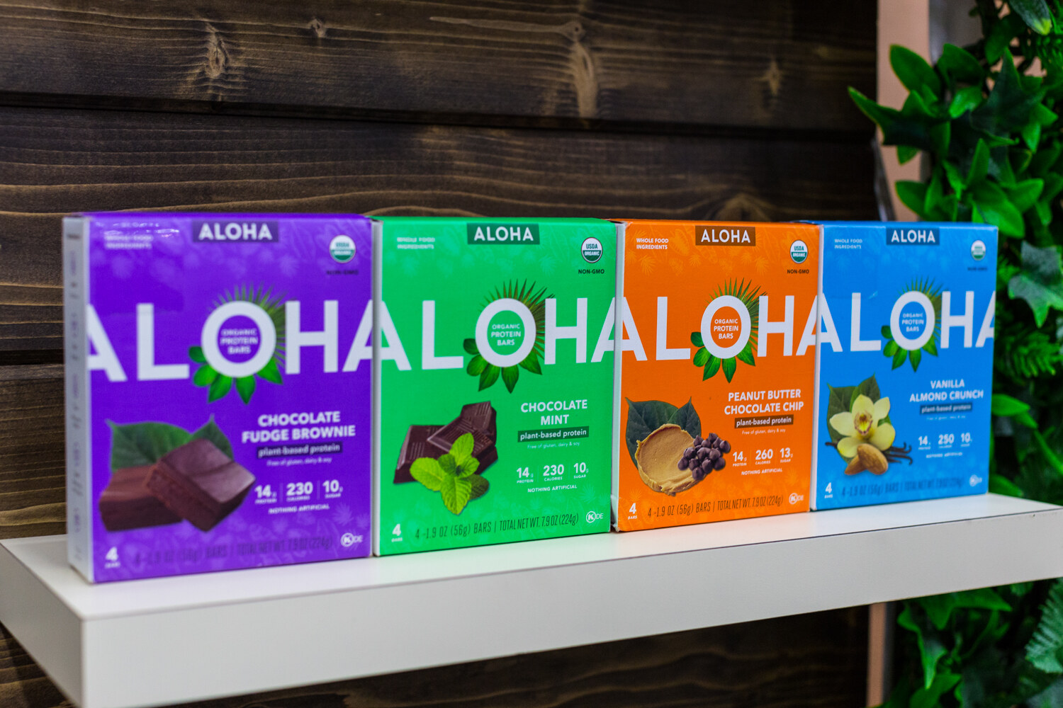Four boxes of Aloha protein bars in purple, green, orange and blue on a white shelf with greenery at the right.