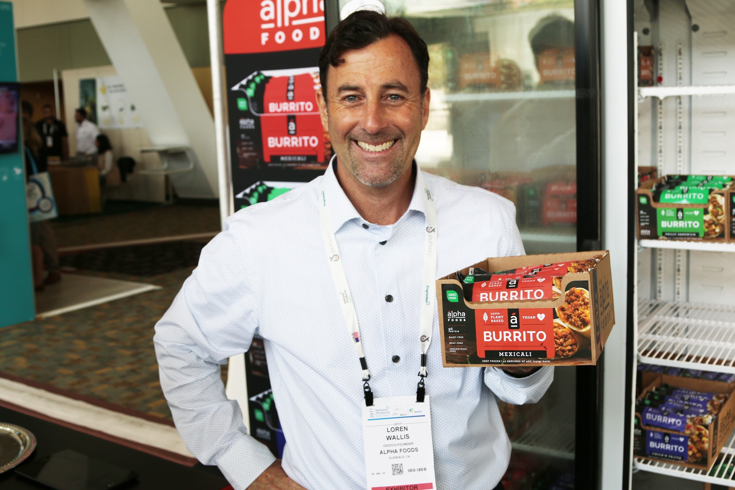 A smiling man with a lanyard holds an open carton of Alpha Foods burritos beside a freezer case, with a carpeted hall behind him.