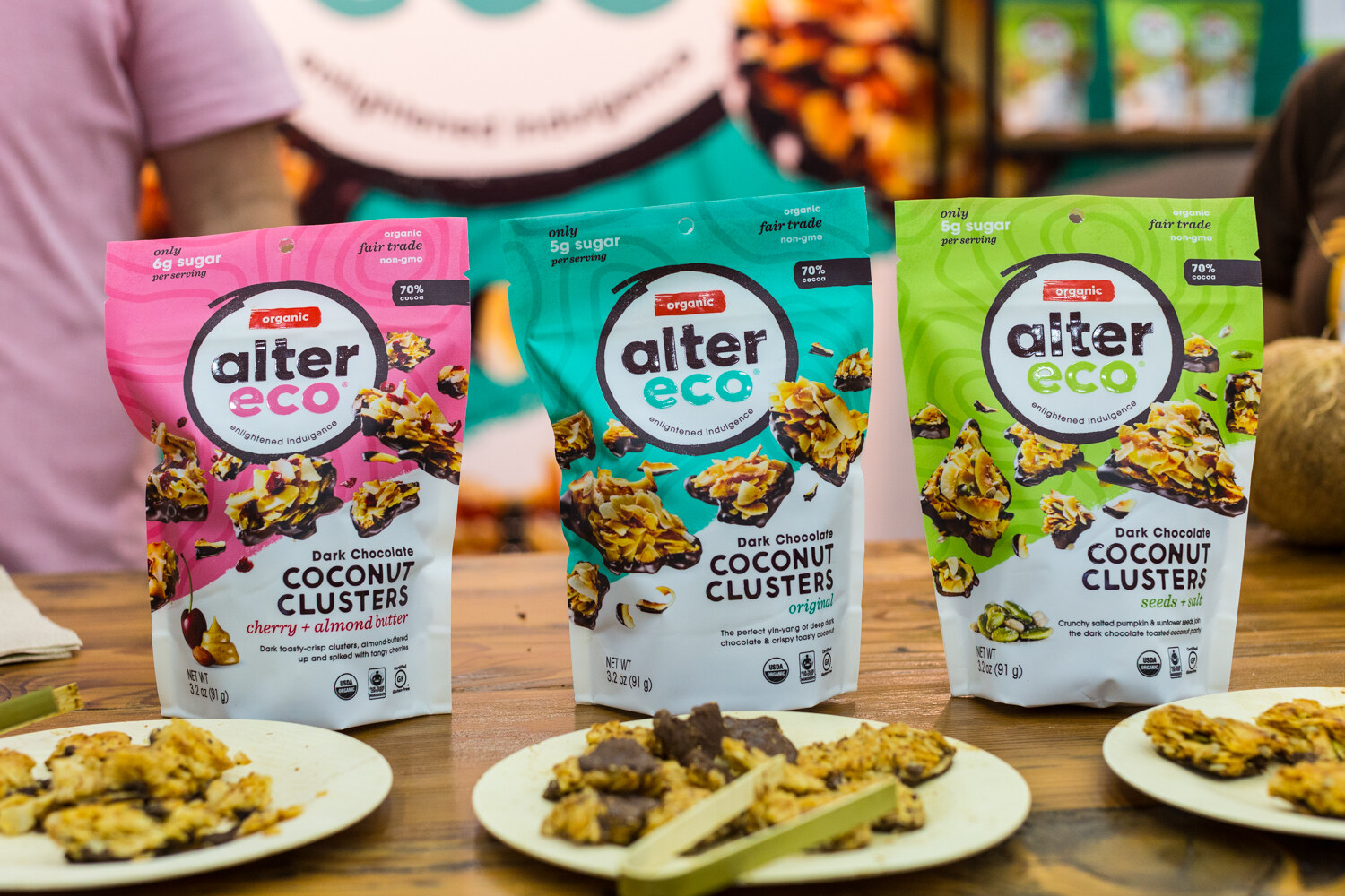 Pink, teal and green Alter Eco coconut clusters pouches in front of a blurred booth worker, with sample plates in front.