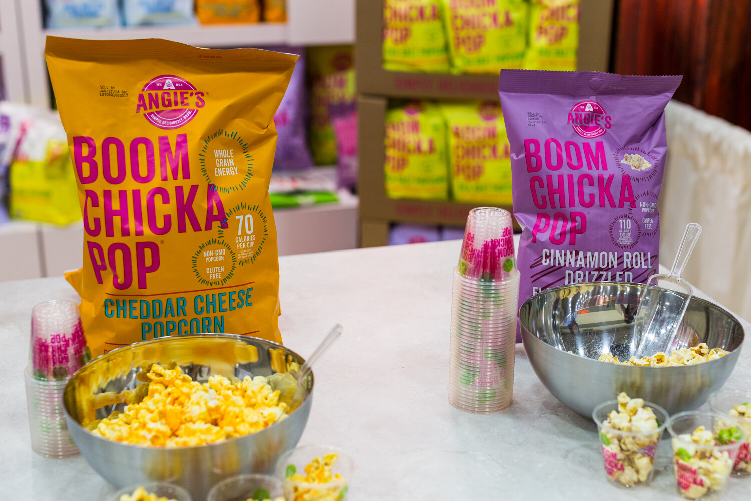 Two large popcorn bags, yellow cheddar and purple cinnamon roll, behind steel bowls of popcorn and small sample cups on a white counter.
