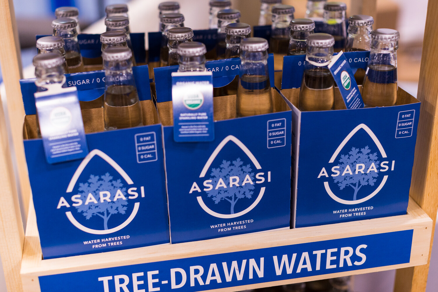 Blue Asarasi four-pack carriers holding glass bottles on a wooden rack above a blue label reading Tree-Drawn Waters.