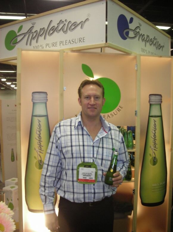 A man in a plaid shirt holds a green Appletiser bottle in a lit wooden booth with Appletiser and Grapetiser signs and large bottle graphics.