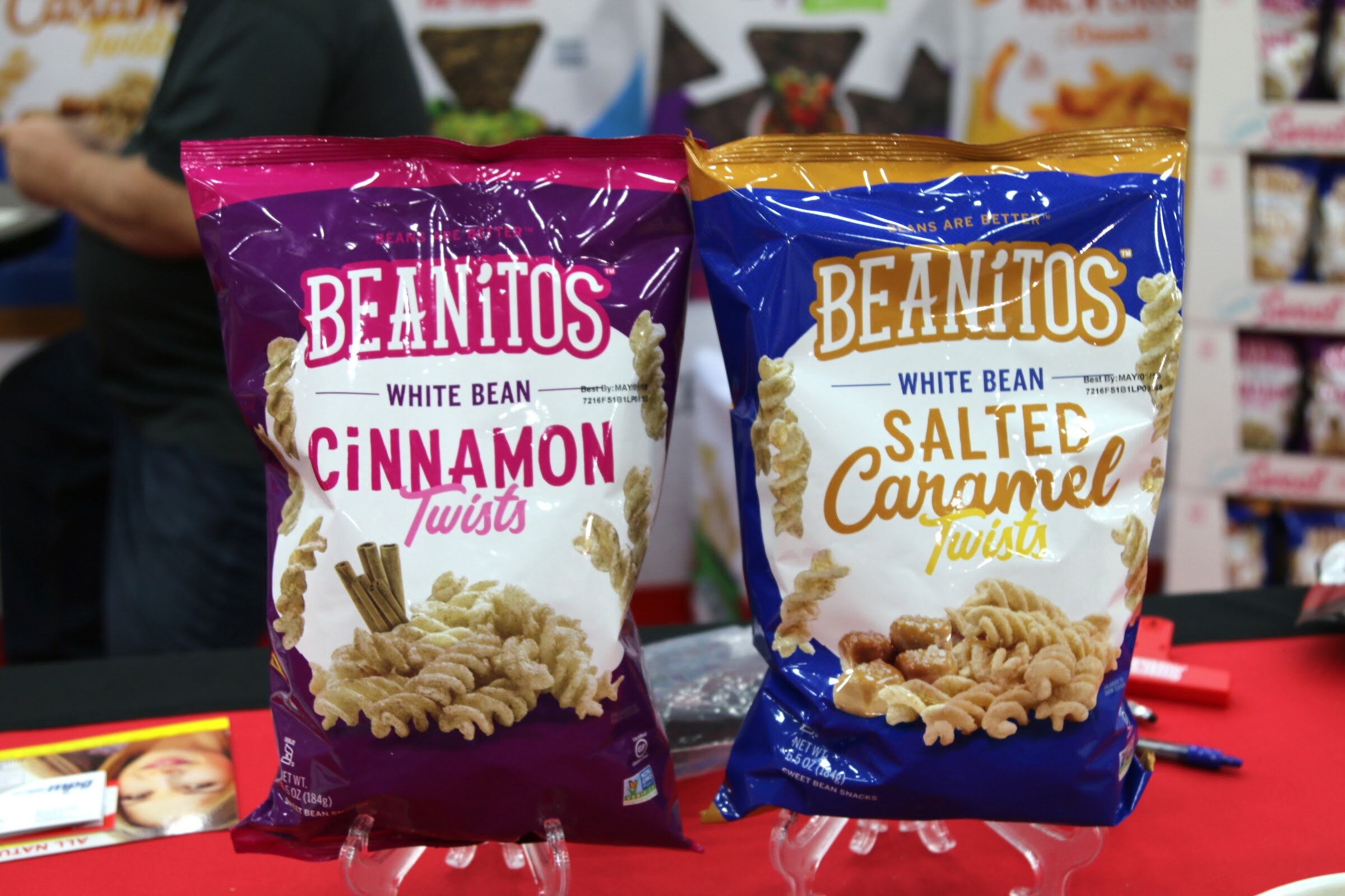 Two large bags of Beanitos white bean twists, one purple and one blue, on a red tablecloth with a booth worker blurred behind.