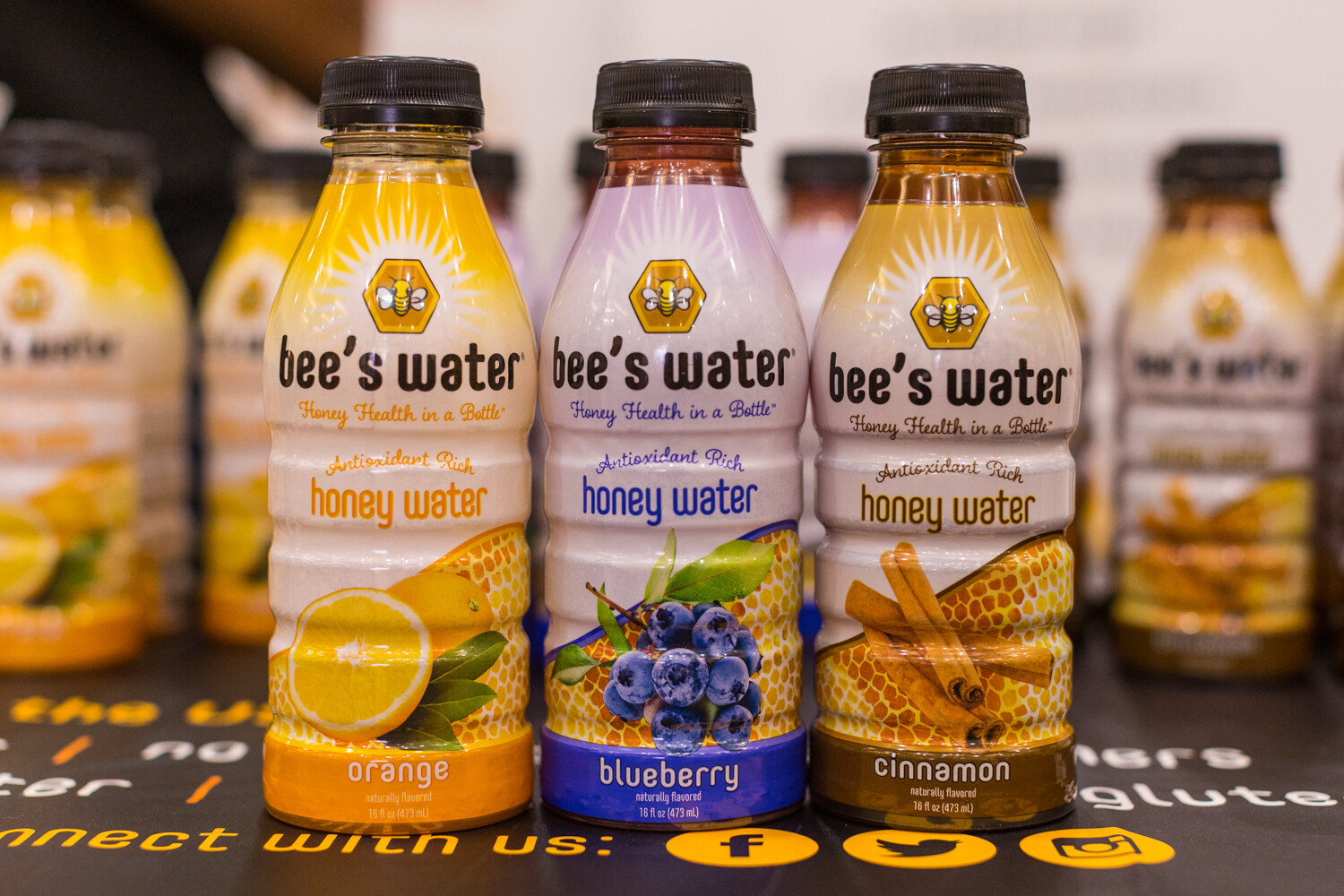 Three bee's water bottles labeled orange, blueberry and cinnamon honey water with more bottles blurred behind.