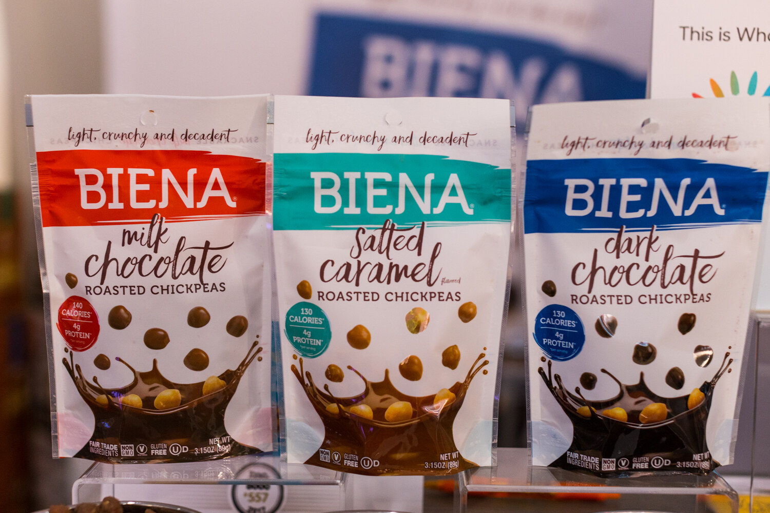 Three Biena roasted chickpea bags with red, teal and blue bands on clear acrylic stands, with part of a NEXTY sign at the edge.