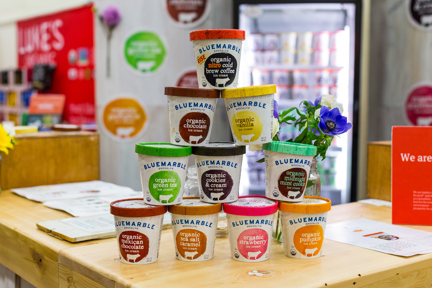 Ten Bluemarble ice cream pints in assorted flavors stacked in a pyramid on a wooden counter with purple flowers and a freezer behind.