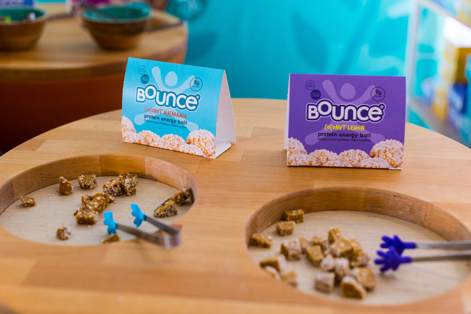 Two Bounce table tents, blue and purple, above round wooden sample trays with cut energy balls and blue and purple tongs.