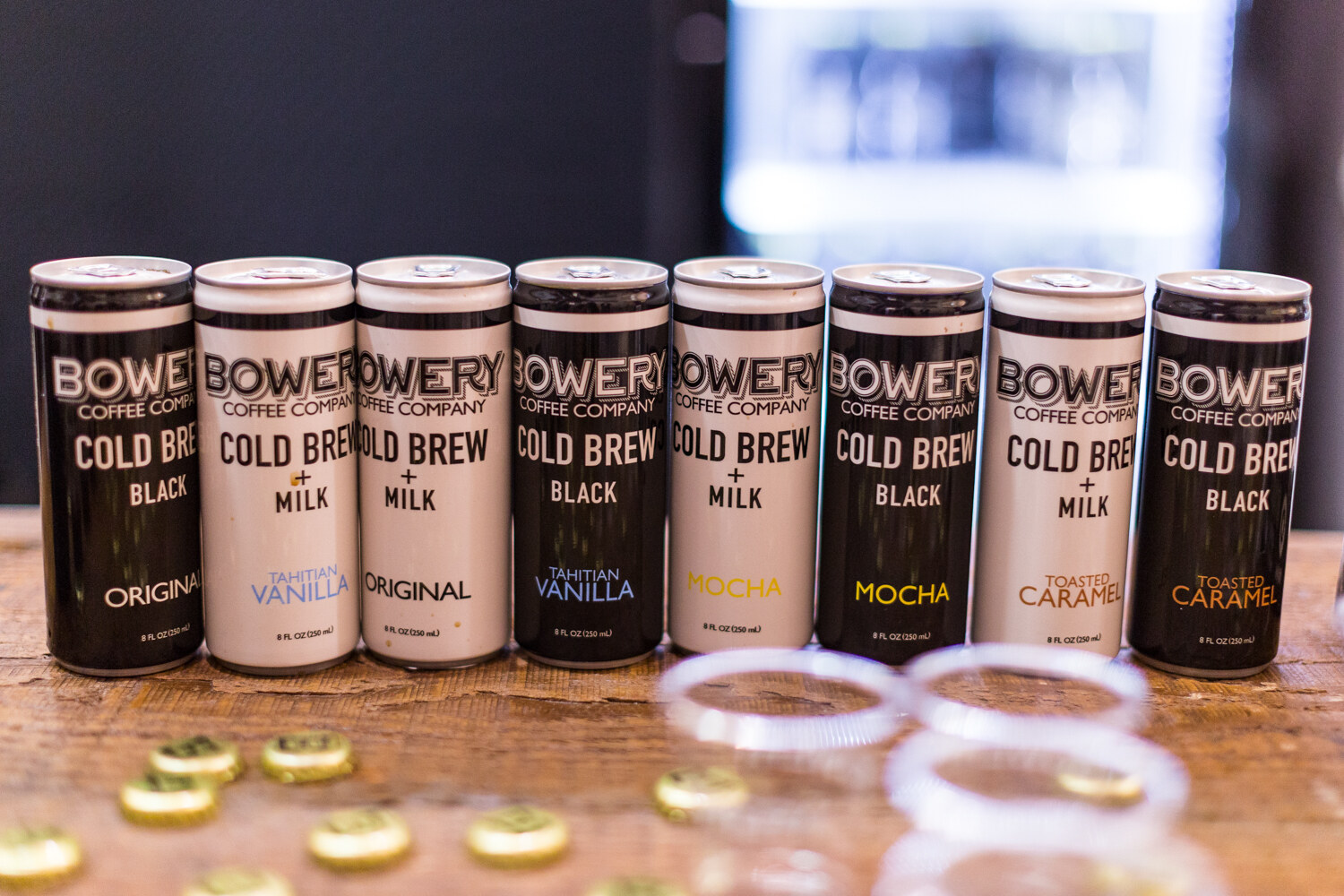 A row of black and cream Bowery Coffee Company cold brew cans in several flavors, with sample cups and bottle caps in front.