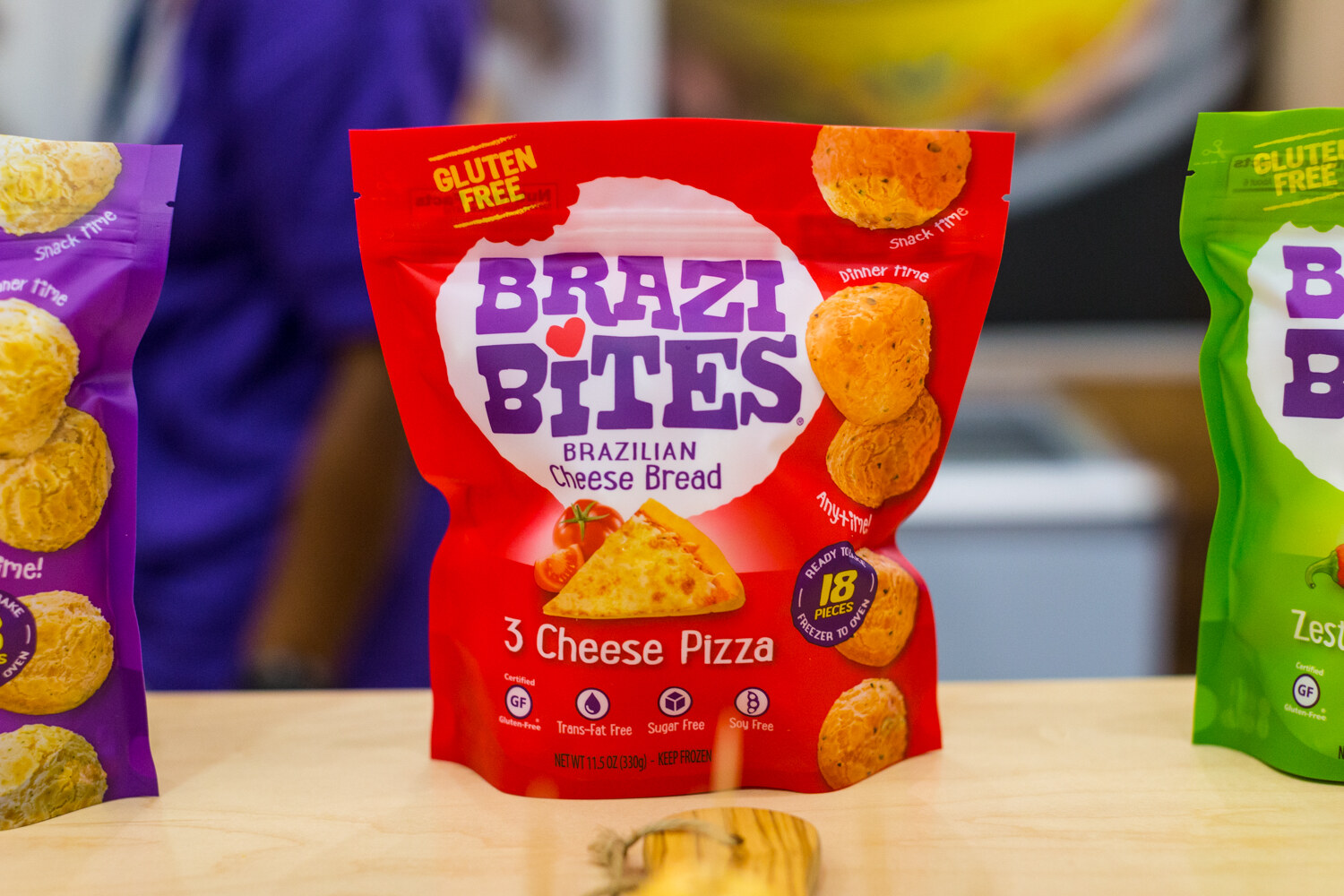 A red Brazi Bites 3 Cheese Pizza bag in sharp focus with purple and green bags at its sides.