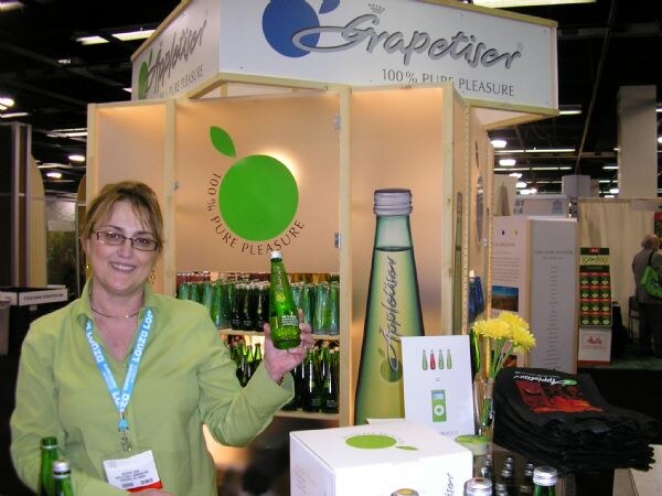 A smiling woman in a green shirt and glasses holds up an Appletiser bottle at the Grapetiser and Appletiser booth, with bottles on shelves and a box on the counter.