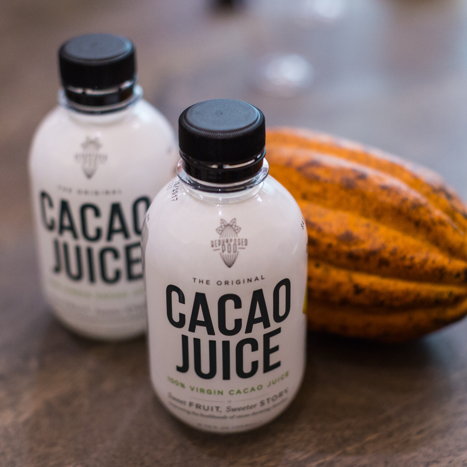 Two white bottles labeled The Original Cacao Juice next to an orange-yellow cacao pod, shot at close range.