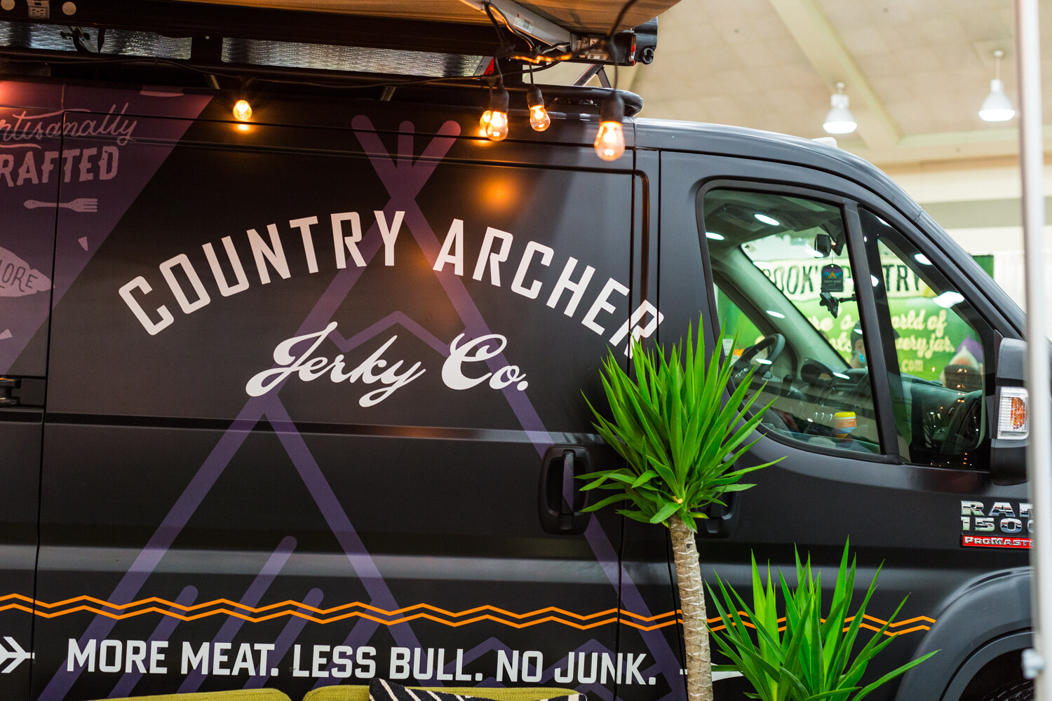 The side of a dark van lettered Country Archer Jerky Co. with string lights, a potted plant and the slogan More Meat. Less Bull. No Junk.
