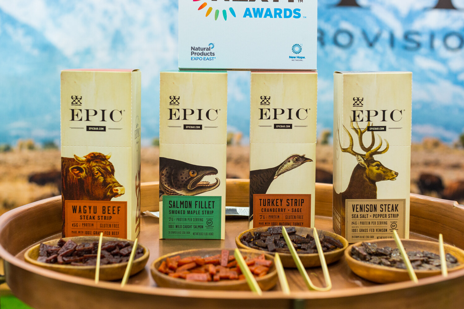 Four cream Epic boxes with animal illustrations on a wooden tray with small bowls of jerky, a NEXTY Awards card above them.