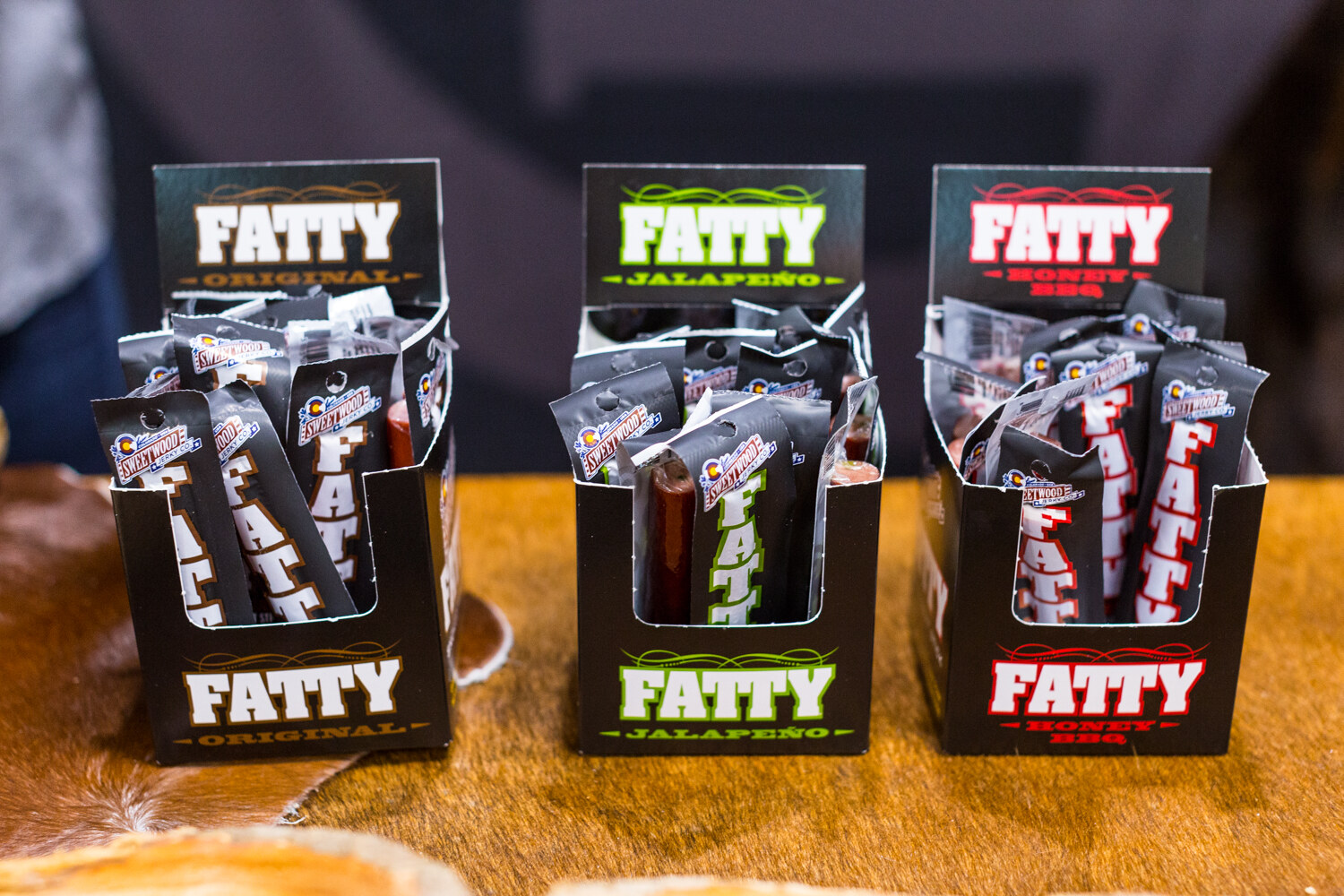 Three black Fatty boxes holding Sweetwood meat stick packs, labeled Original, Jalapeno and Honey BBQ.