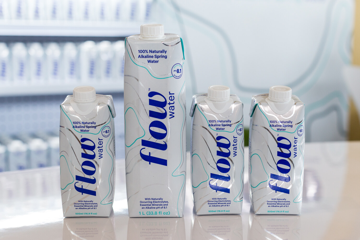 Four white Flow alkaline spring water cartons with blue lettering, one tall liter carton among three smaller ones.