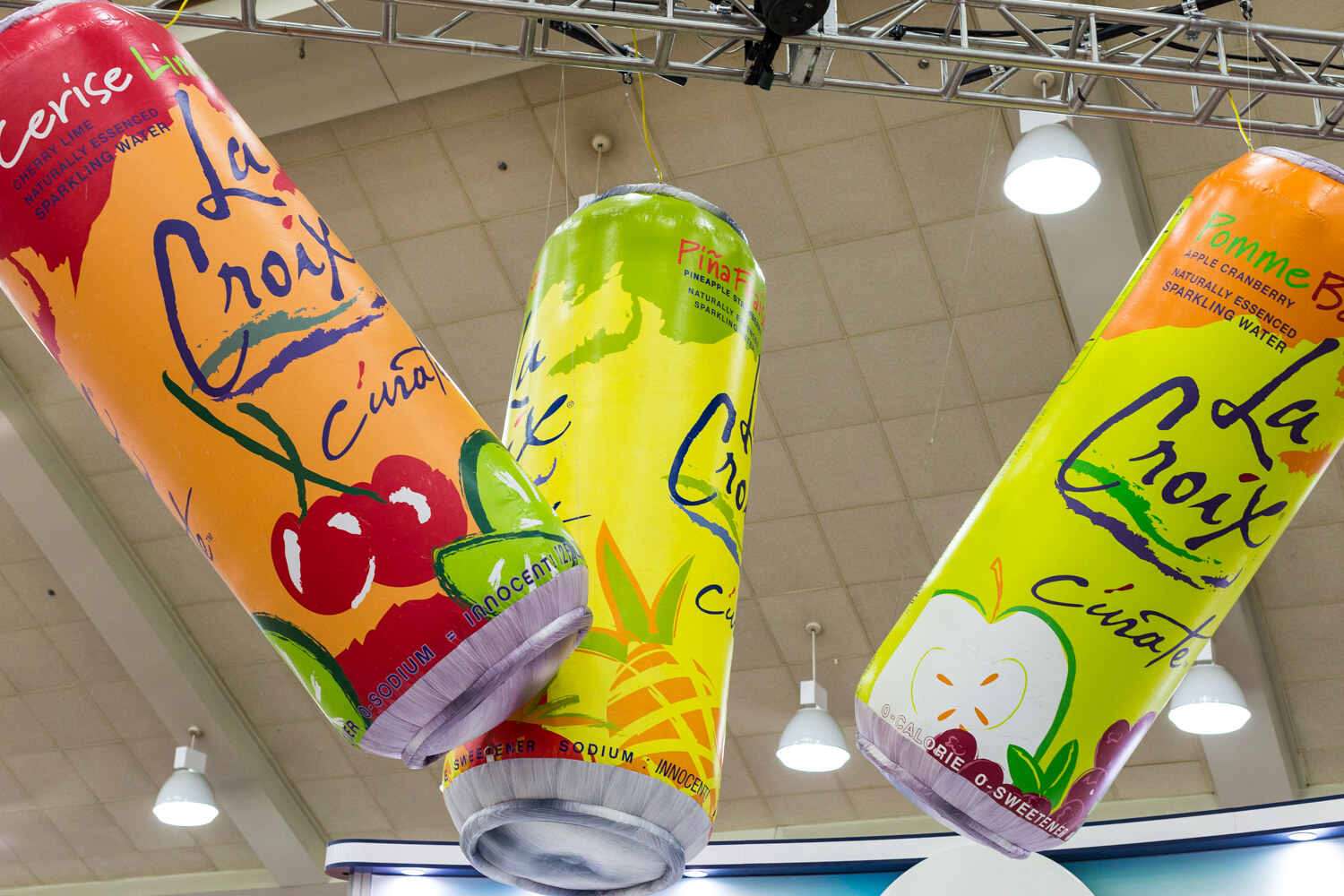 Three giant inflatable LaCroix sparkling water cans hanging from a ceiling truss with hall lights around them.