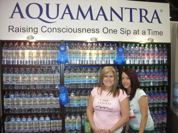 Two women stand in front of shelves of Aquamantra water bottles with blue ribbons, under a sign reading Raising Consciousness One Sip at a Time. One wears a pink I Am Loved shirt.