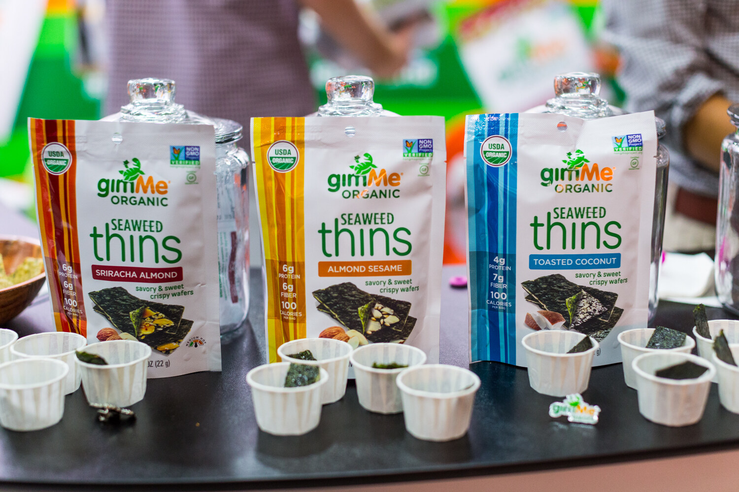 Three gimMe Organic Seaweed Thins bags in red, orange and blue stripes behind small paper cups with seaweed sheets.