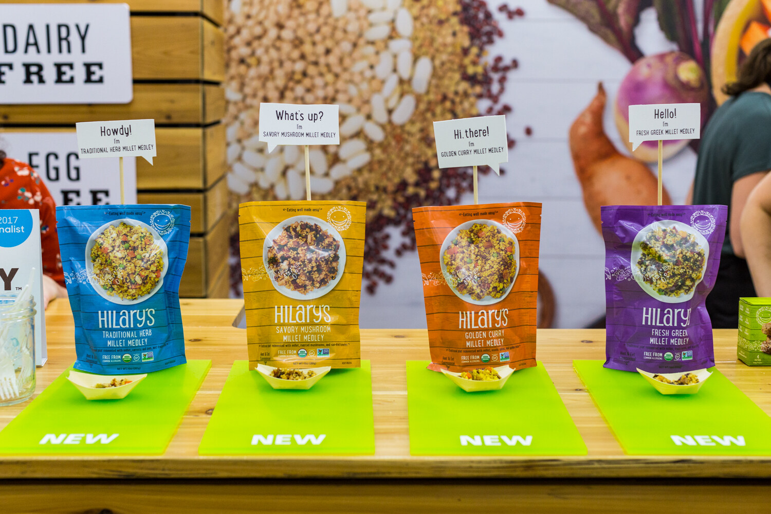Blue, yellow, orange and purple Hilary's millet medley pouches on a wooden counter with green mats printed NEW and small sample boats.