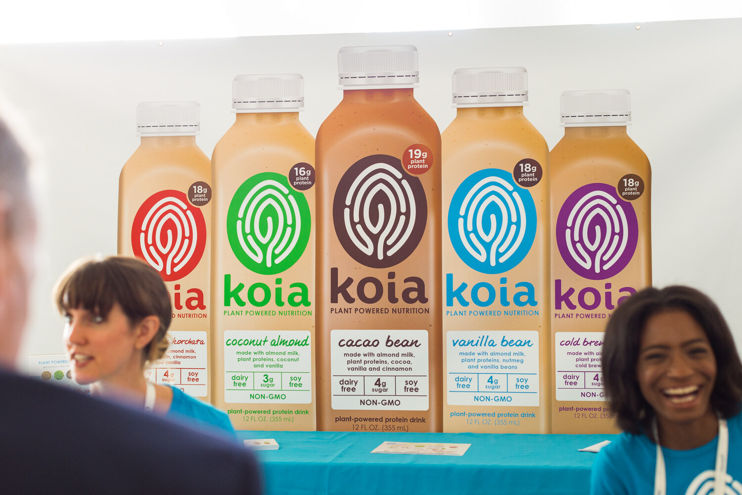Two women in teal shirts at a teal-draped table in front of a large banner of five Koia protein drink bottles in different flavors.