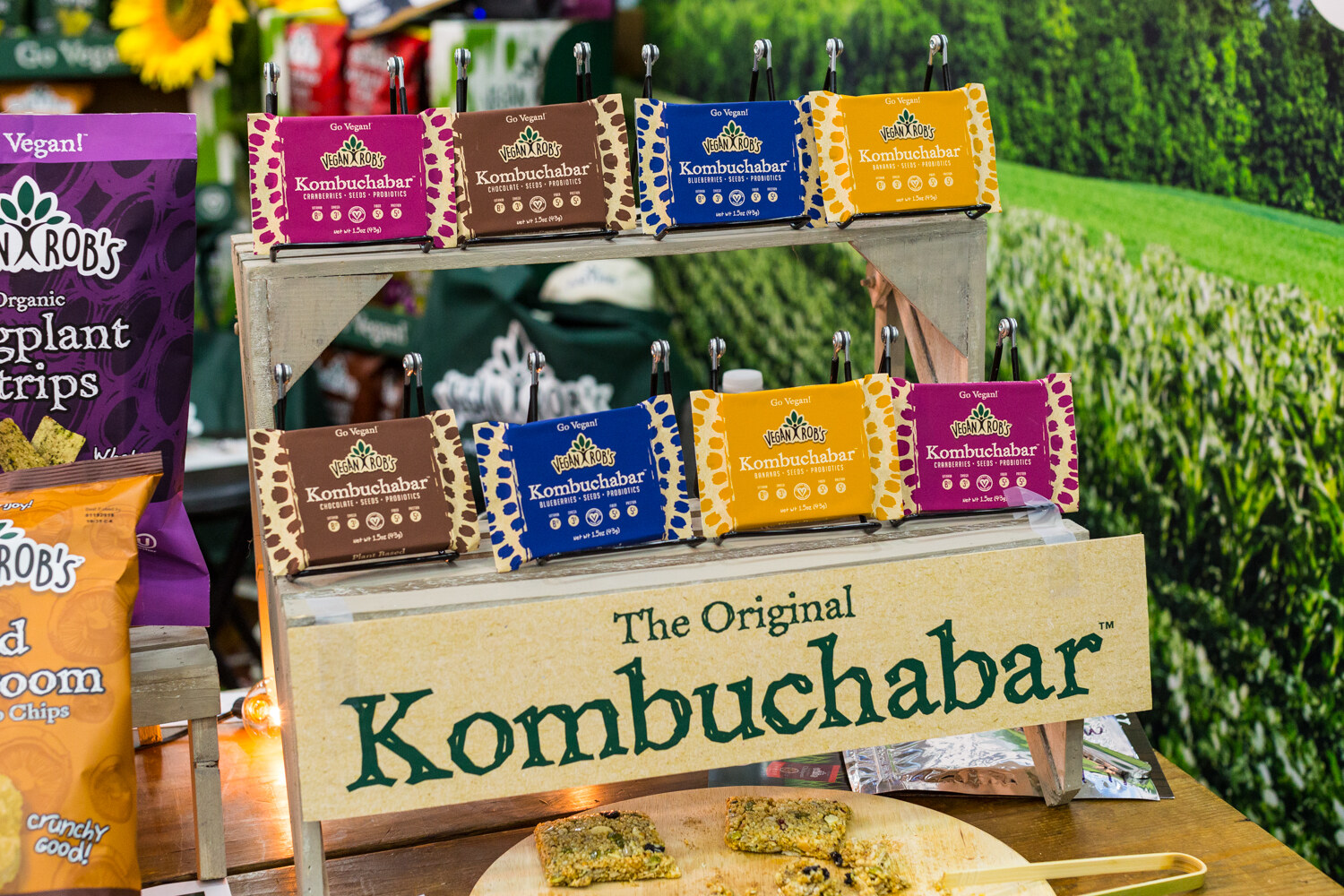 Two rows of colorful Kombuchabar packages clipped to a wooden stand labeled The Original Kombuchabar, with a green field backdrop.