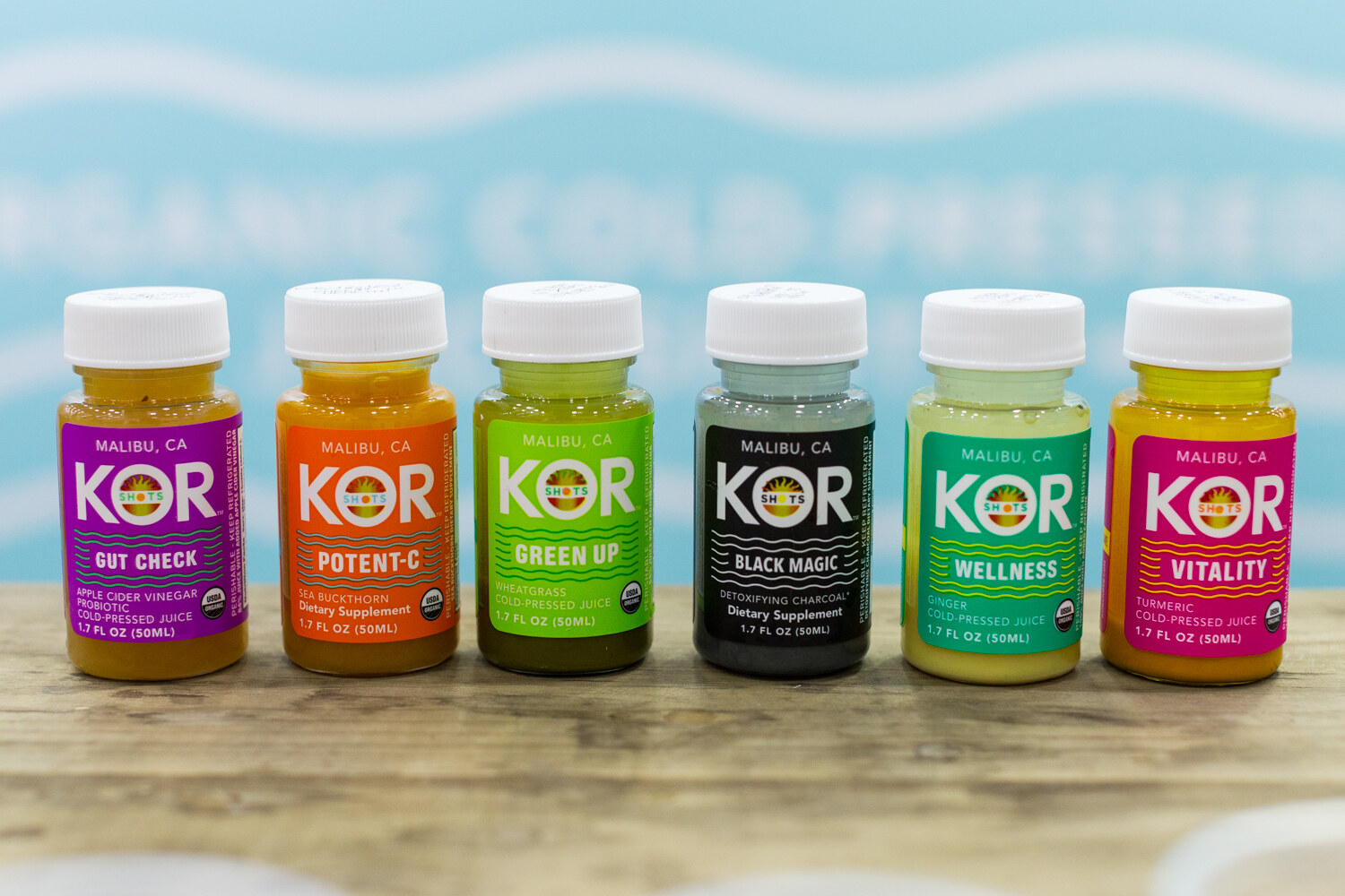 Six small colorful KOR Shots juice bottles in a row on a rustic wooden counter against a pale blue backdrop.