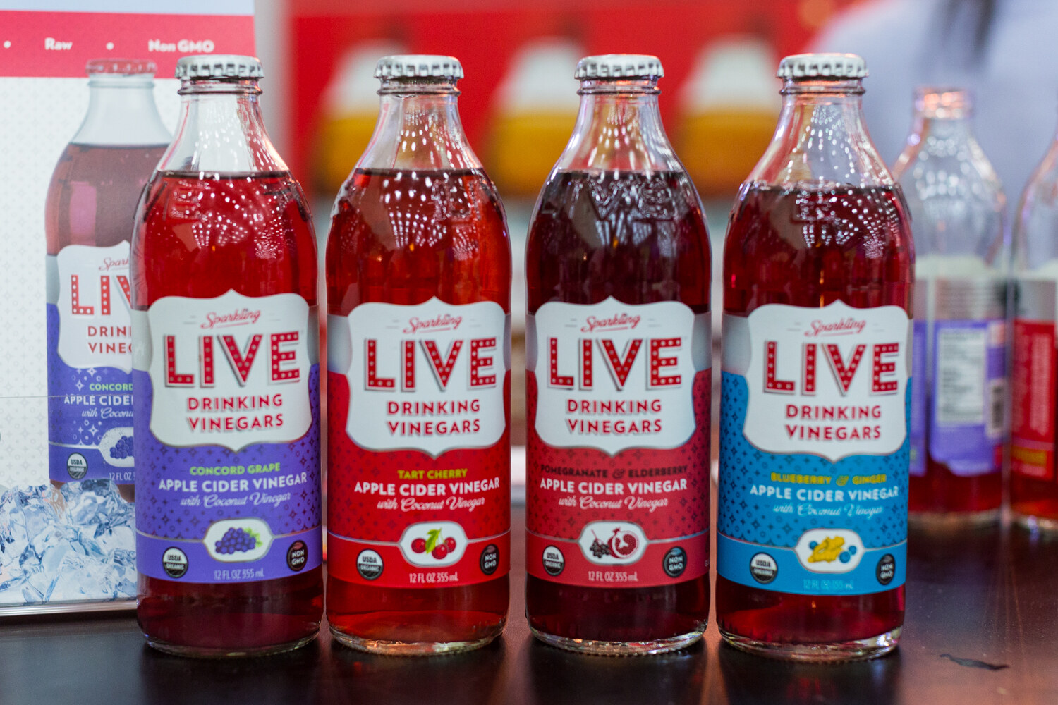 Four glass bottles of LIVE Drinking Vinegars with white labels in purple, red and blue bands.