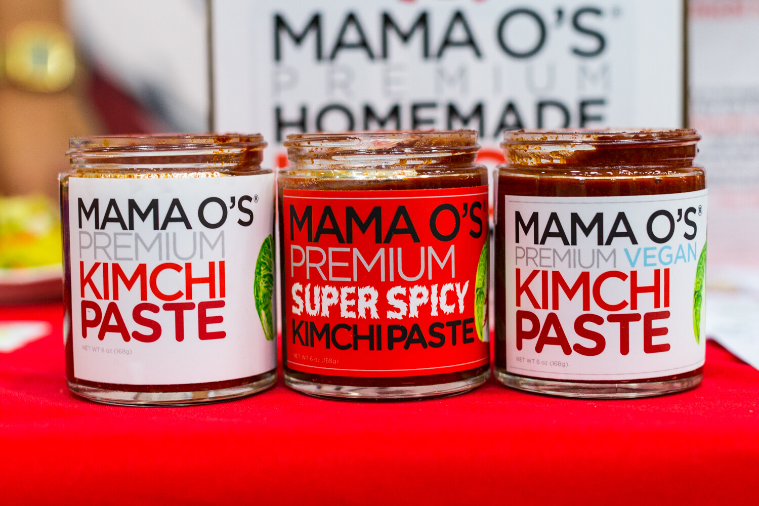 Three jars labeled Mama O's Premium Kimchi Paste on a red cloth in front of a Mama O's Premium Homemade sign.
