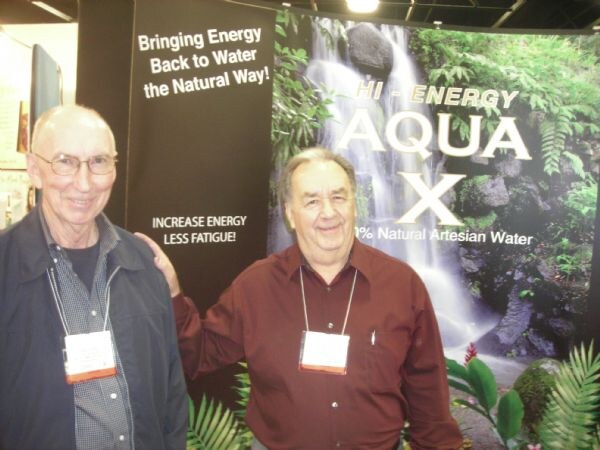 Two older men with badges stand together in front of an Aqua X banner showing a waterfall and the text Bringing Energy Back to Water the Natural Way.