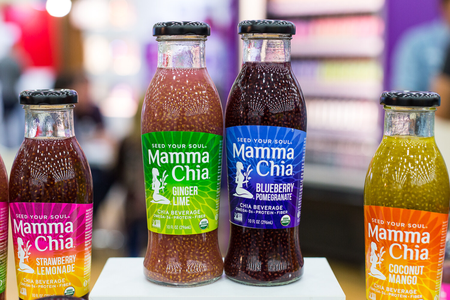 Four glass bottles of Mamma Chia chia beverage with colorful labels in front of a blurred exhibit hall.