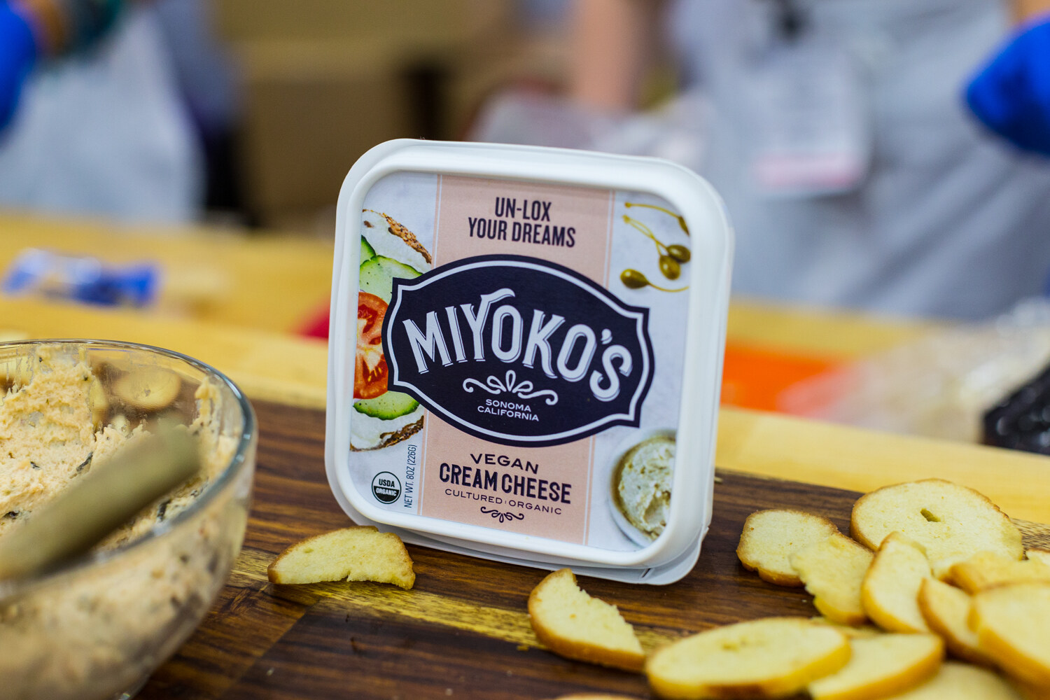 A white Miyoko's Vegan Cream Cheese tub upright on a wood board with toasted bagel chips and a glass bowl of spread beside it.