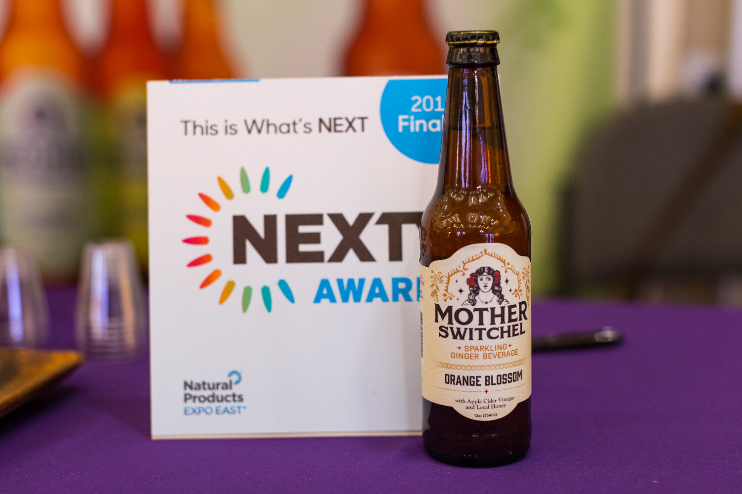 A brown bottle of Mother Switchel Orange Blossom beside a card reading This is What's NEXT, NEXTY Awards, Natural Products Expo East.