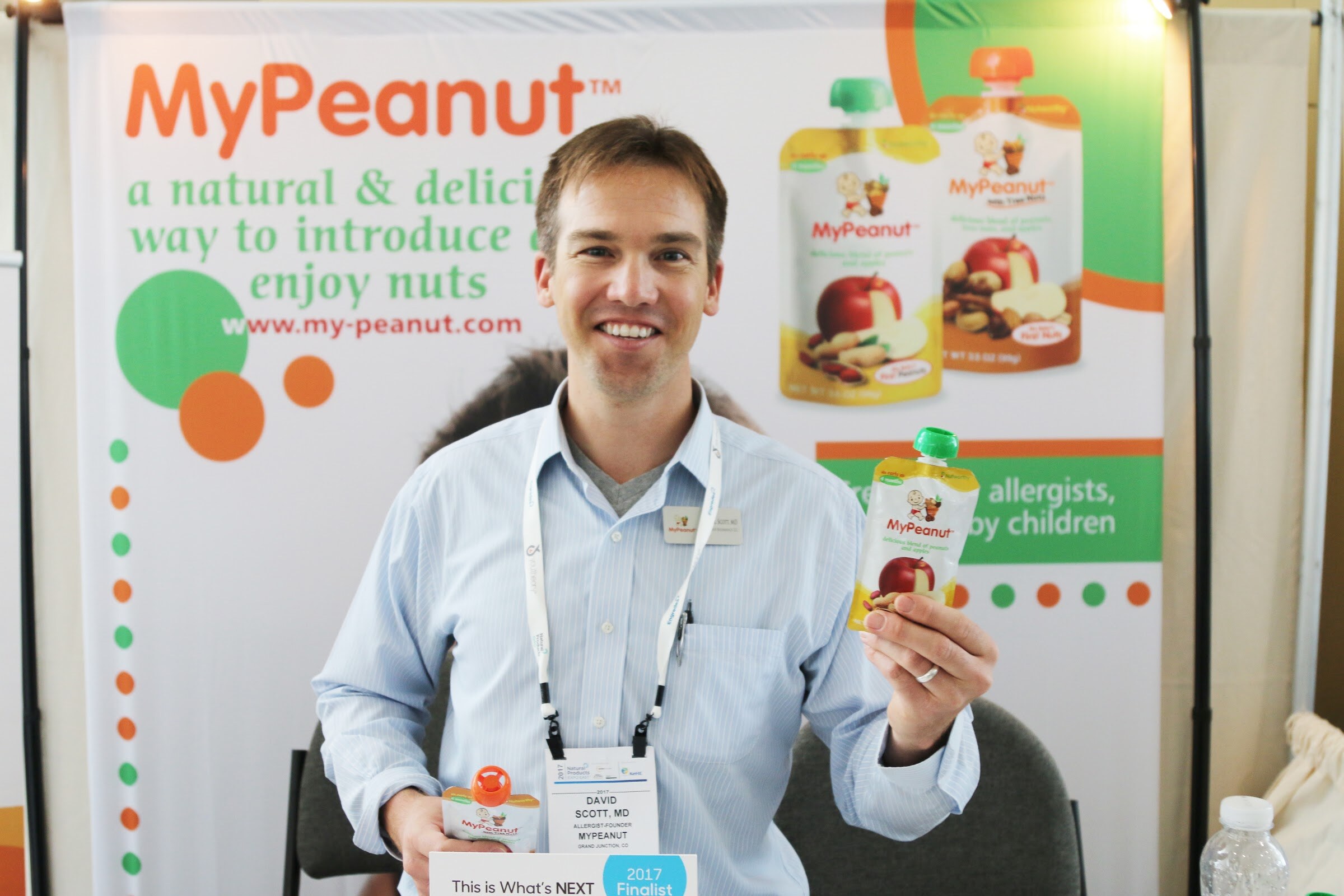 A smiling man in a light blue shirt and lanyard holds a MyPeanut pouch in front of a MyPeanut banner.