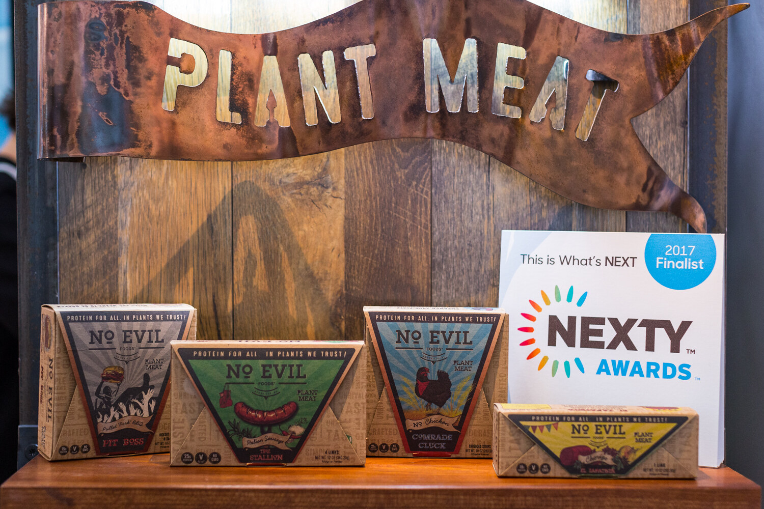 Four triangular No Evil Foods boxes on a wooden shelf under a copper Plant Meat sign, beside a 2017 NEXTY Awards Finalist card.