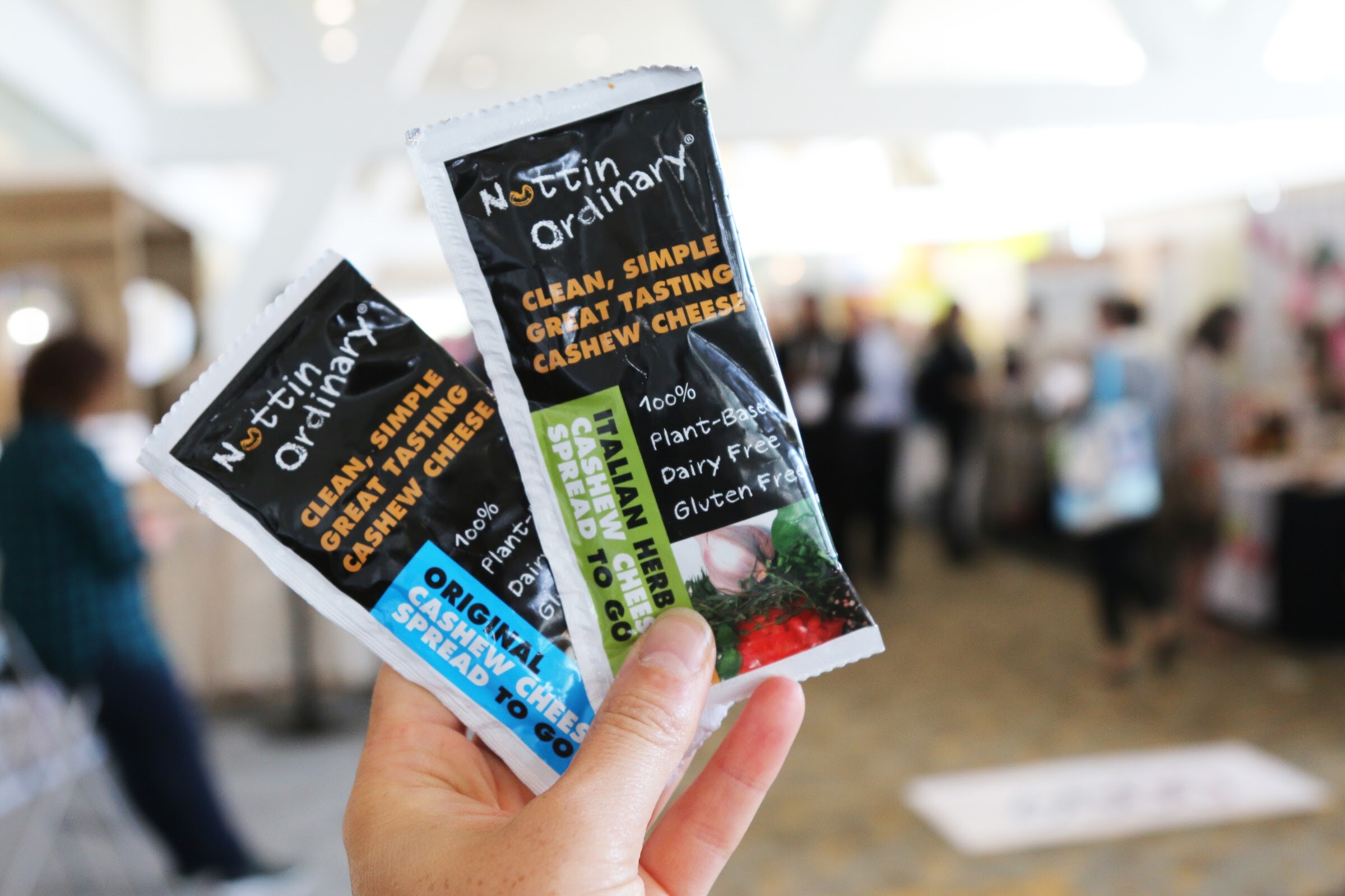 A hand holds two black and orange cashew cheese spread packets in front of a blurred crowd on an exhibit hall floor.