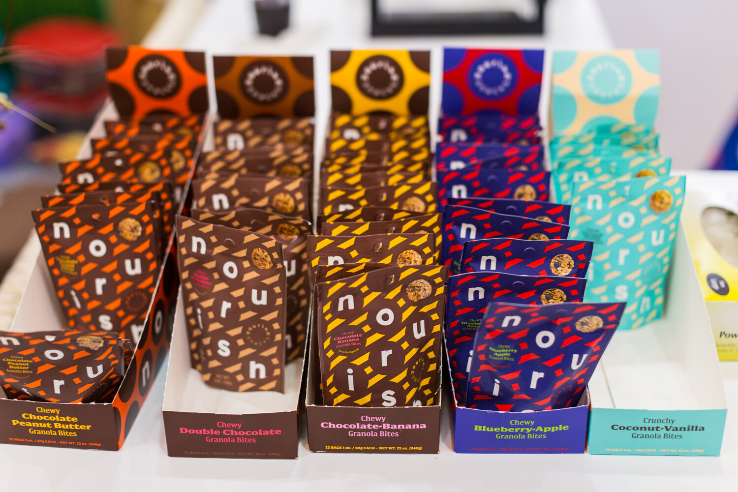 Five open cardboard boxes of colorful patterned Nourish granola bite pouches in orange, brown, yellow, blue and teal.