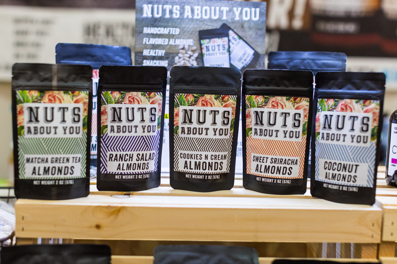 Five black Nuts About You pouches with floral labels, from matcha green tea to coconut almonds, on a light wood shelf.