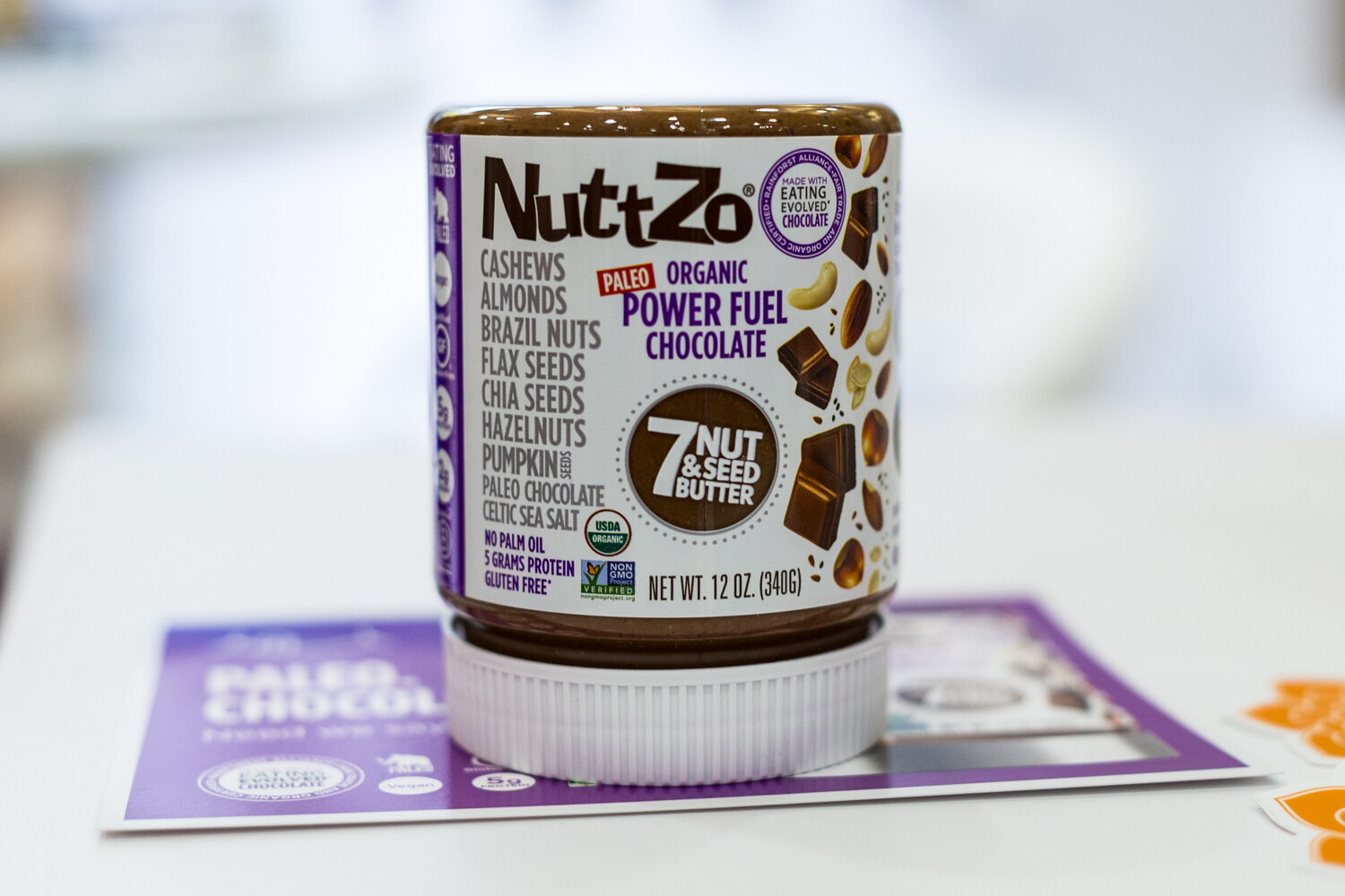 A single NuttZo jar listing its seven nuts and seeds, balanced upside down on a white counter with a purple card beneath.