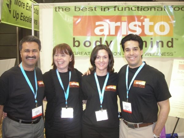 Four people in black Aristo body + mind shirts and blue lanyards stand close together smiling in front of the green Aristo body + mind booth banner.