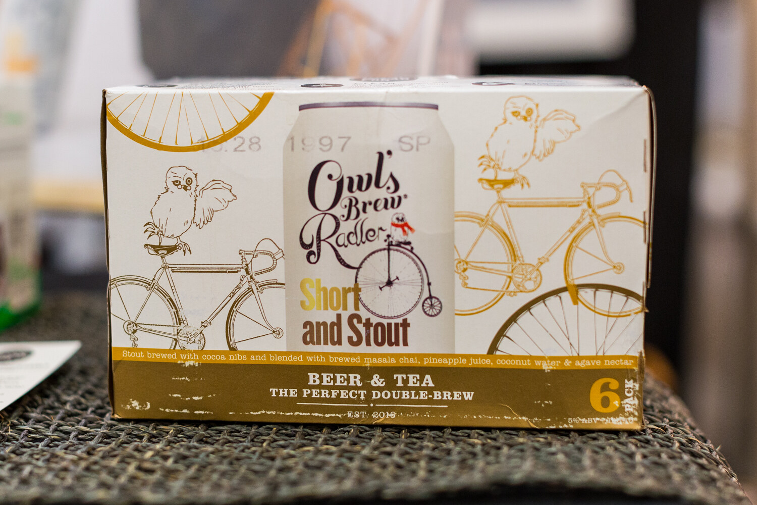 A cream and gold six-pack box with owl and bicycle drawings reading Owl's Brew Radler Short and Stout, Beer and Tea.